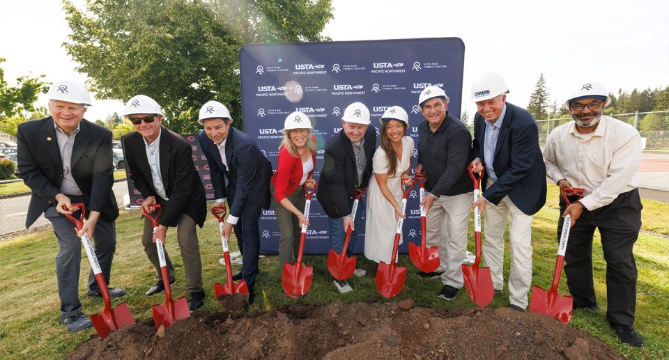 Nine people in hard hats holding red shovels during a groundbreaking ceremony, standing in front of a sign outdoors.