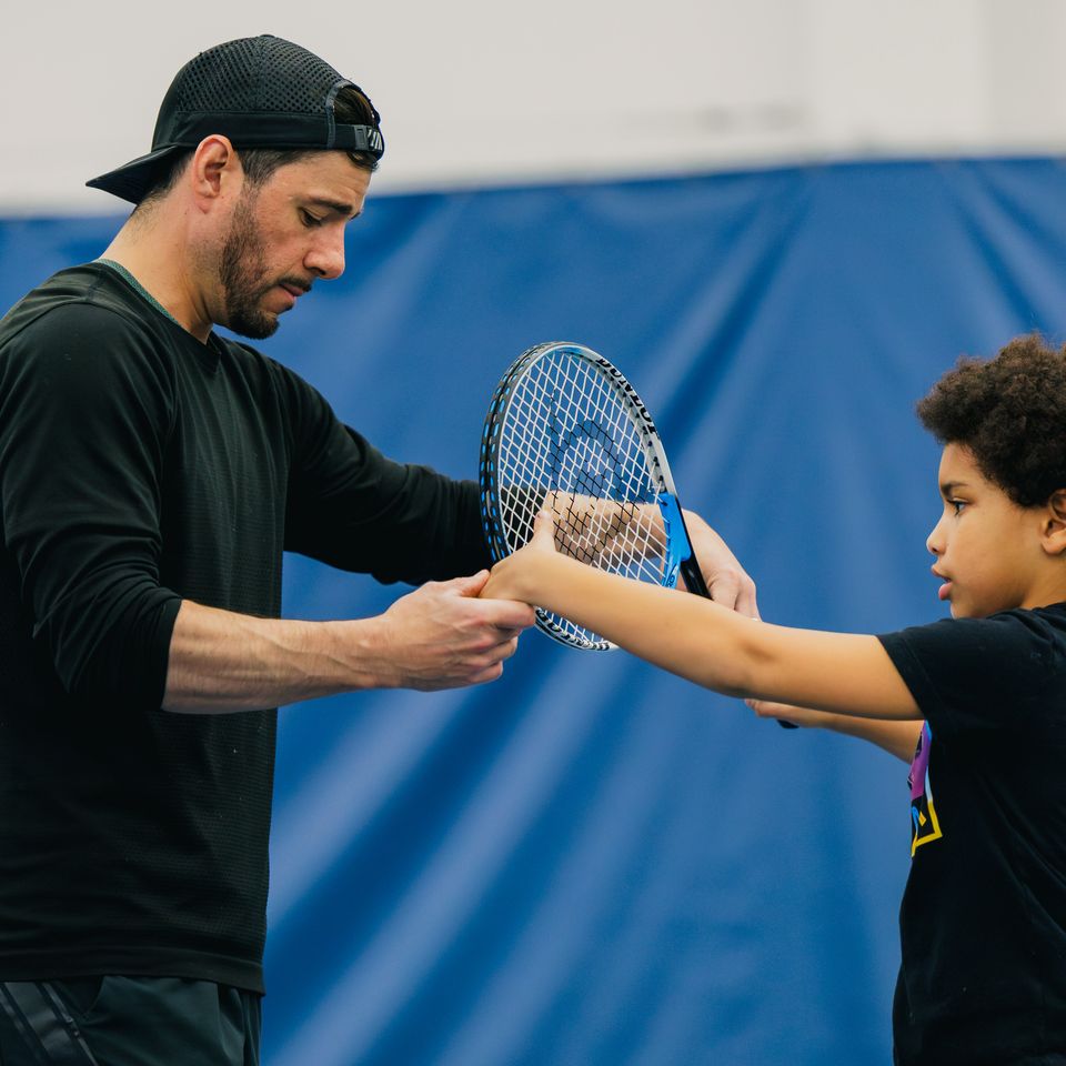 A tennis coach in a black cap instructs a young child on tennis racket grip indoors, against a blue background.