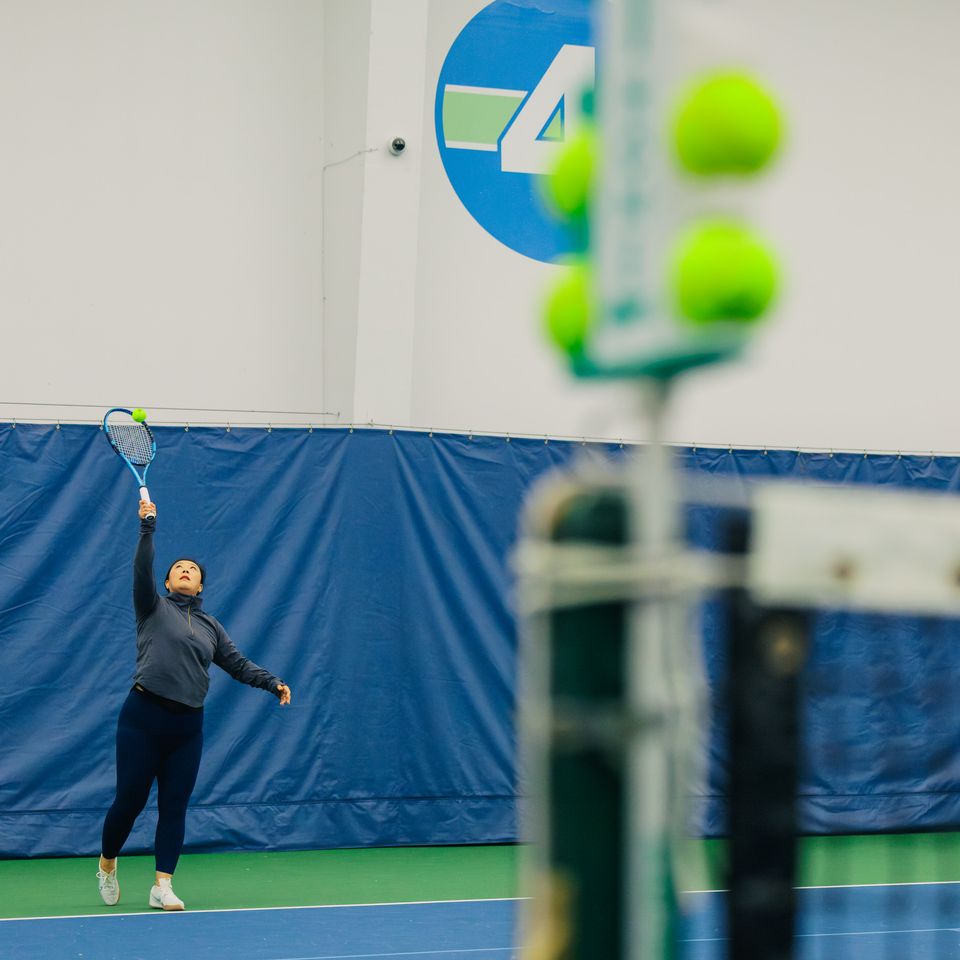 Person serving a tennis ball indoors, with a focus on their upward swing and multiple balls visible on the right.