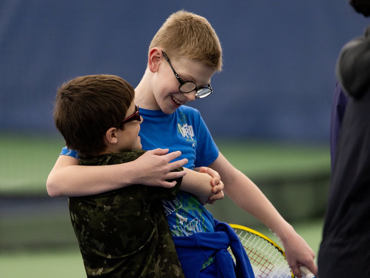 Two boys wearing glasses hug happily on a tennis court. One wears a blue shirt, holding a racket, while the other wears a camouflage shirt.