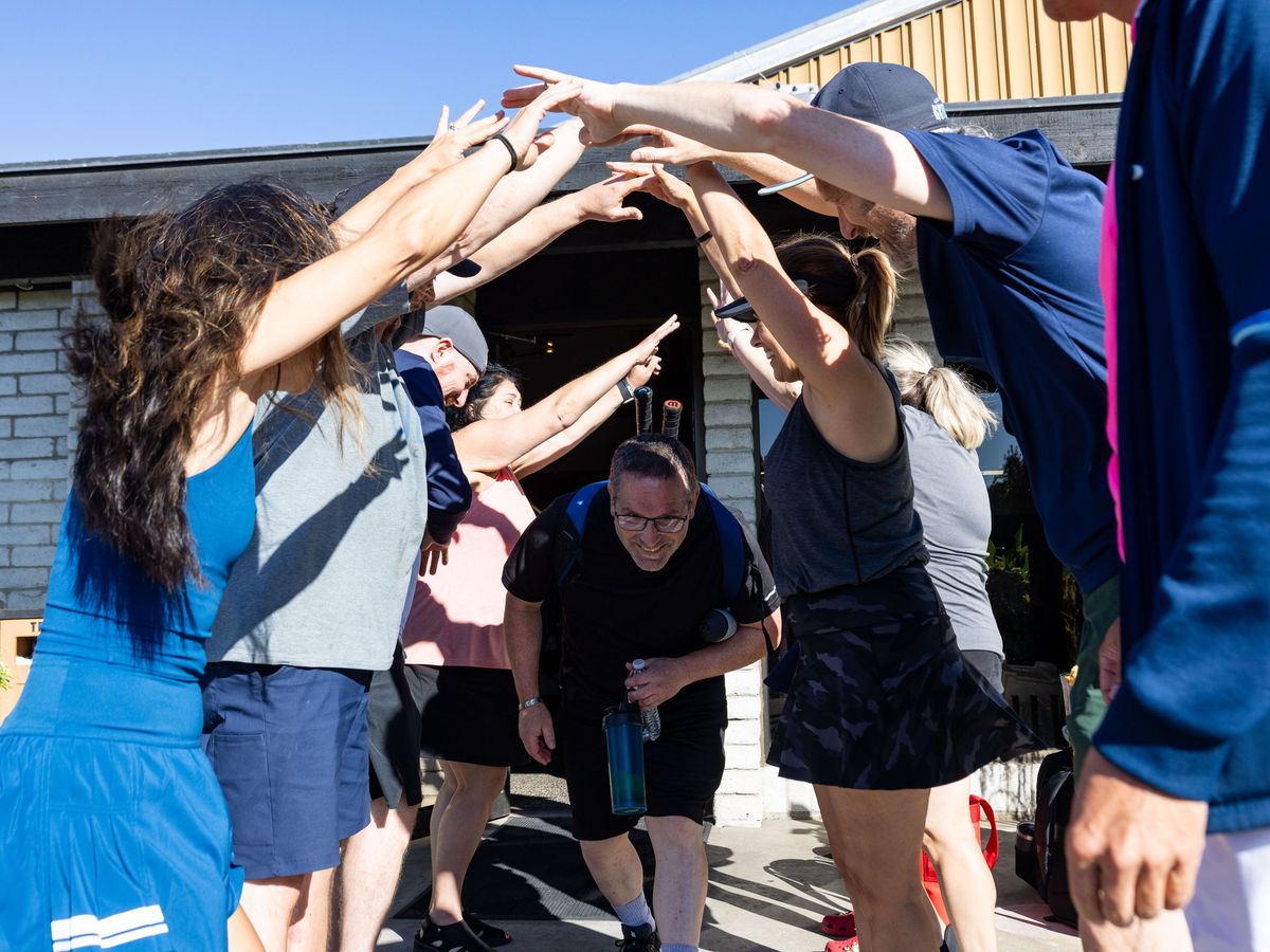 A man with a water bottle ducks under an arch formed by raised arms of smiling people outdoors.