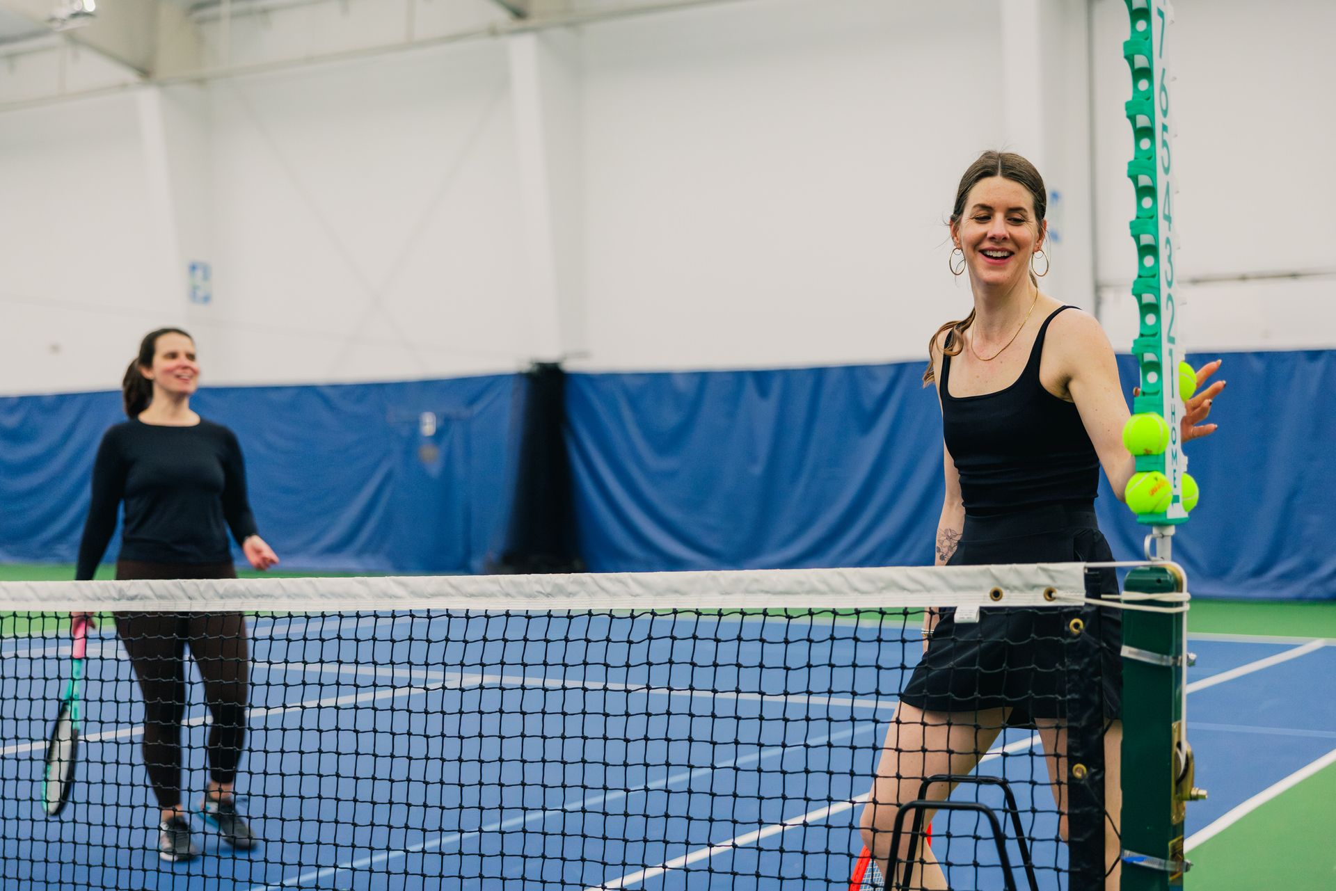Two women on an indoor tennis court, smiling by the net. One holds a racket, the other rests on net post with tennis balls.