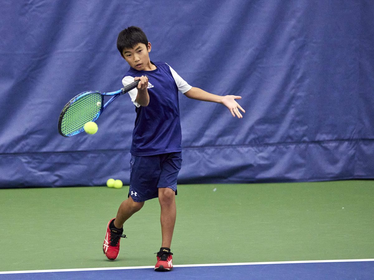 Young tennis player in a navy outfit hits a forehand shot on an indoor court, with tennis balls visible in the background.