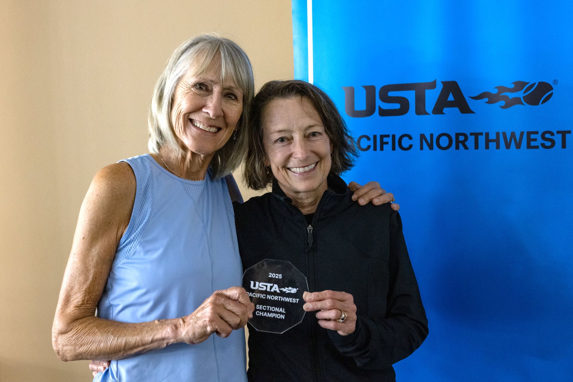 Two smiling women holding a 2025 USTA Pacific Northwest Sectional Champion award, standing in front of a blue banner.