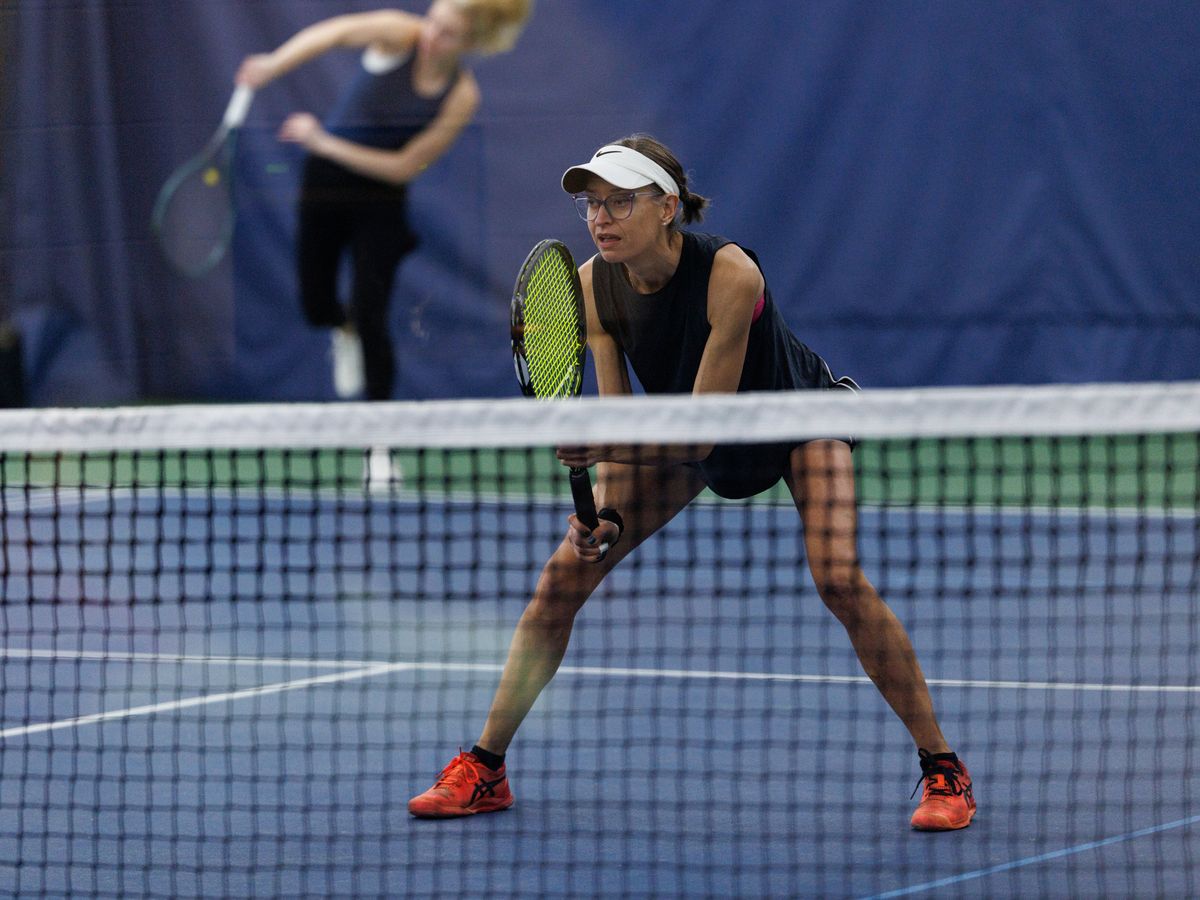 Tennis player in ready position near the net, wearing a visor and glasses, with another player in the background on a blue indoor court.