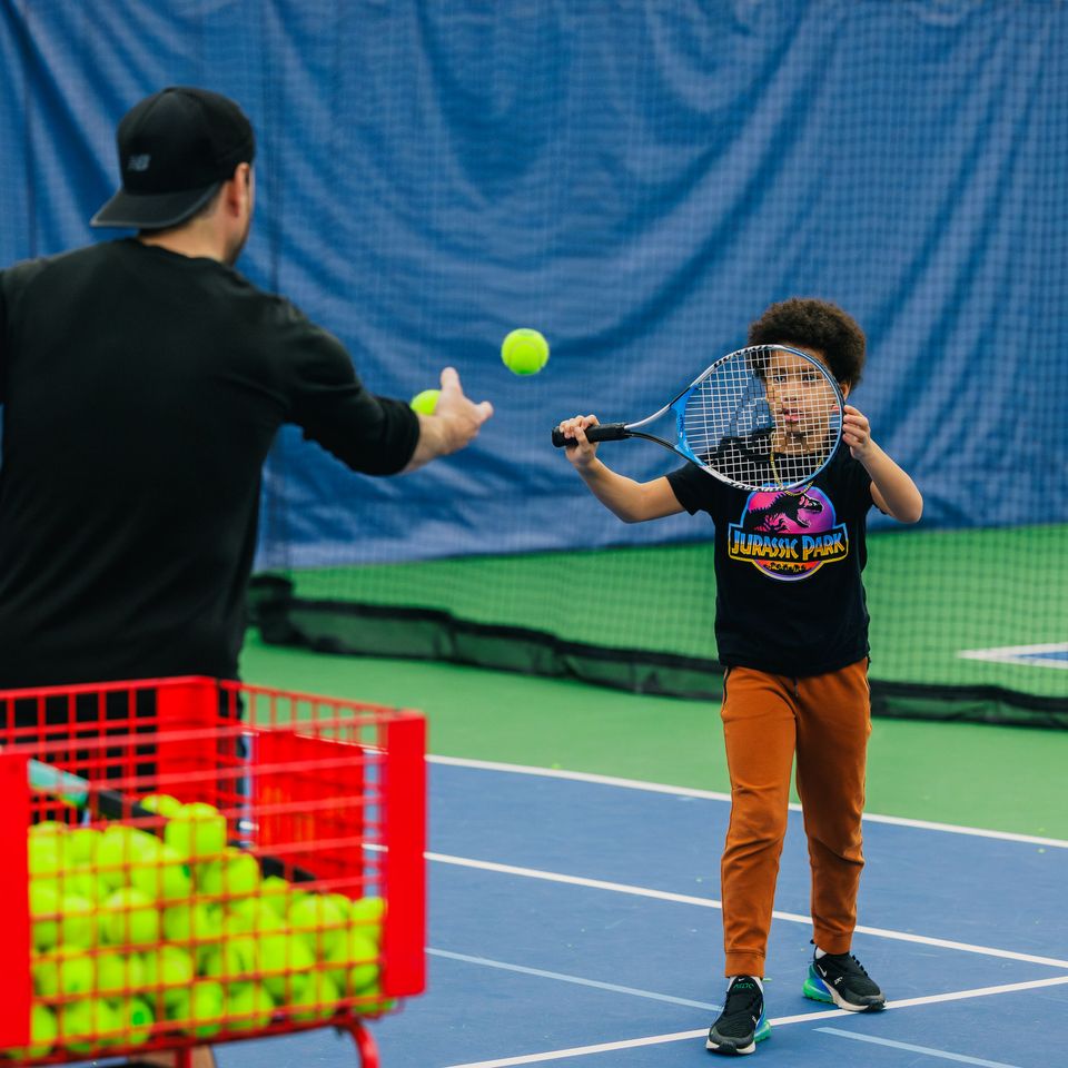 Child practicing tennis with a coach in a blue indoor court, hitting a ball with a racket.