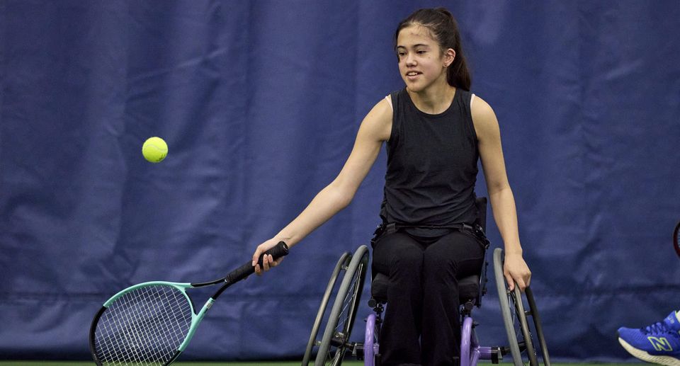 A teen girl n in a wheelchair playing tennis indoors, reaching for a tennis ball with her racket.