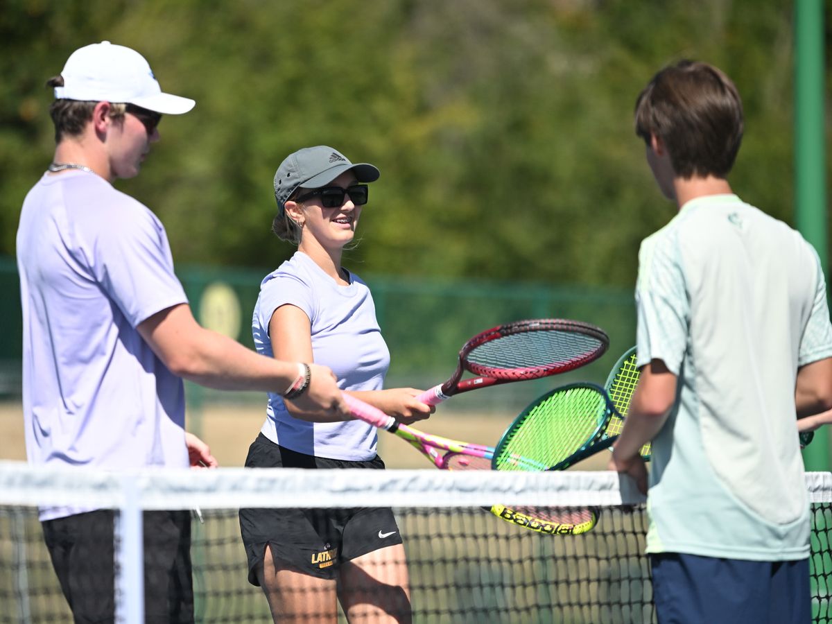 Tennis players in casual attire at the net, exchanging handshakes and smiles, with racquets prominently visible, under sunny skies.