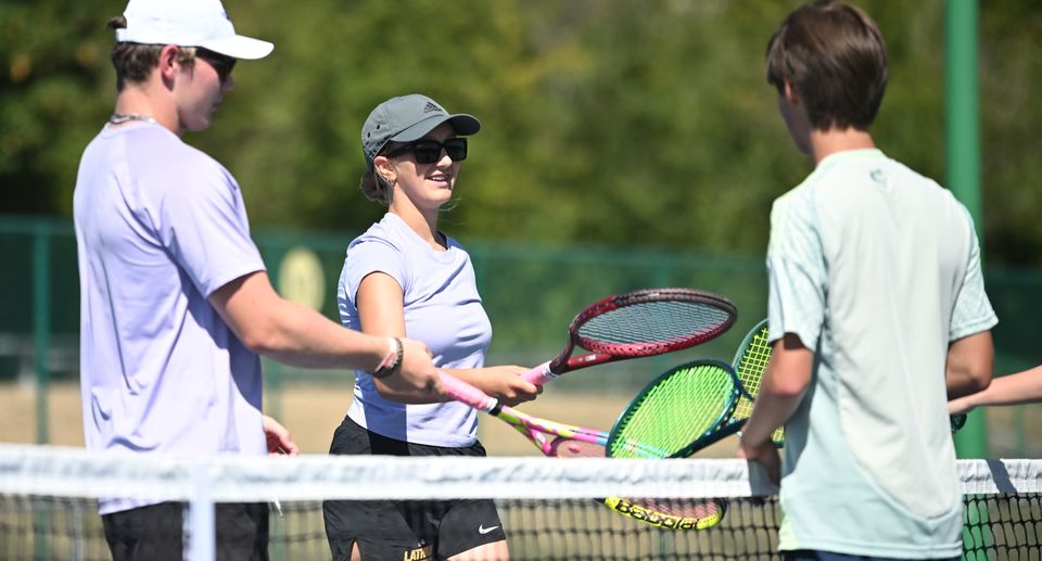 Tennis players in casual attire at the net, exchanging handshakes and smiles, with racquets prominently visible, under sunny skies.