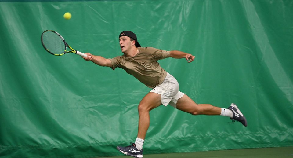 Tennis player in motion, stretching to hit a tennis ball with a racket on an indoor green court, wearing a cap, brown shirt, and white shorts.