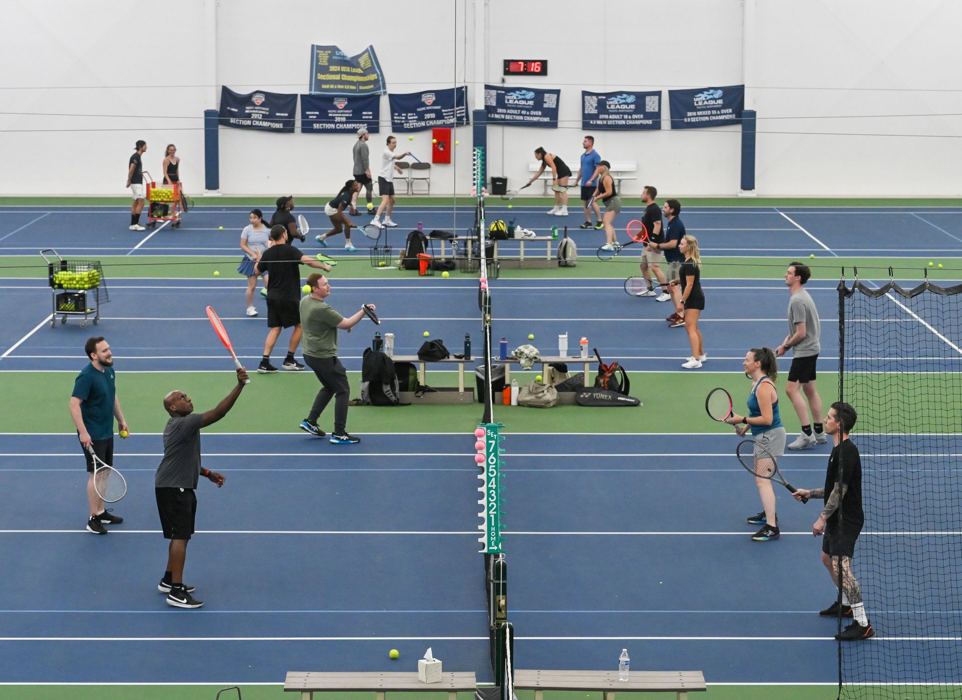 Indoor tennis facility with multiple players practicing on blue and green courts, surrounded by equipment and banners on the wall.