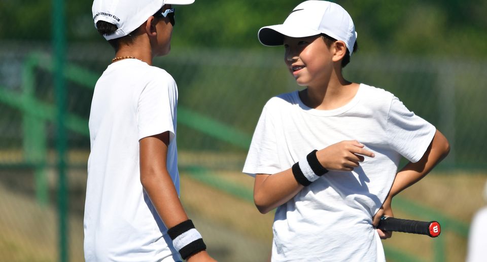 Two boys in white shirts and caps chat during a tennis match, holding rackets and wearing wristbands on a sunny day.