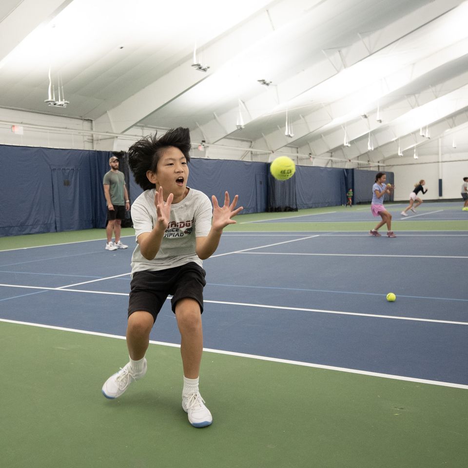 Child eagerly catching a tennis ball on an indoor court with other children practicing in the background.