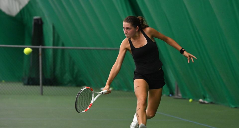 A woman in black sportswear plays tennis indoors, swiftly returning a ball with her racquet, green court and curtain background.