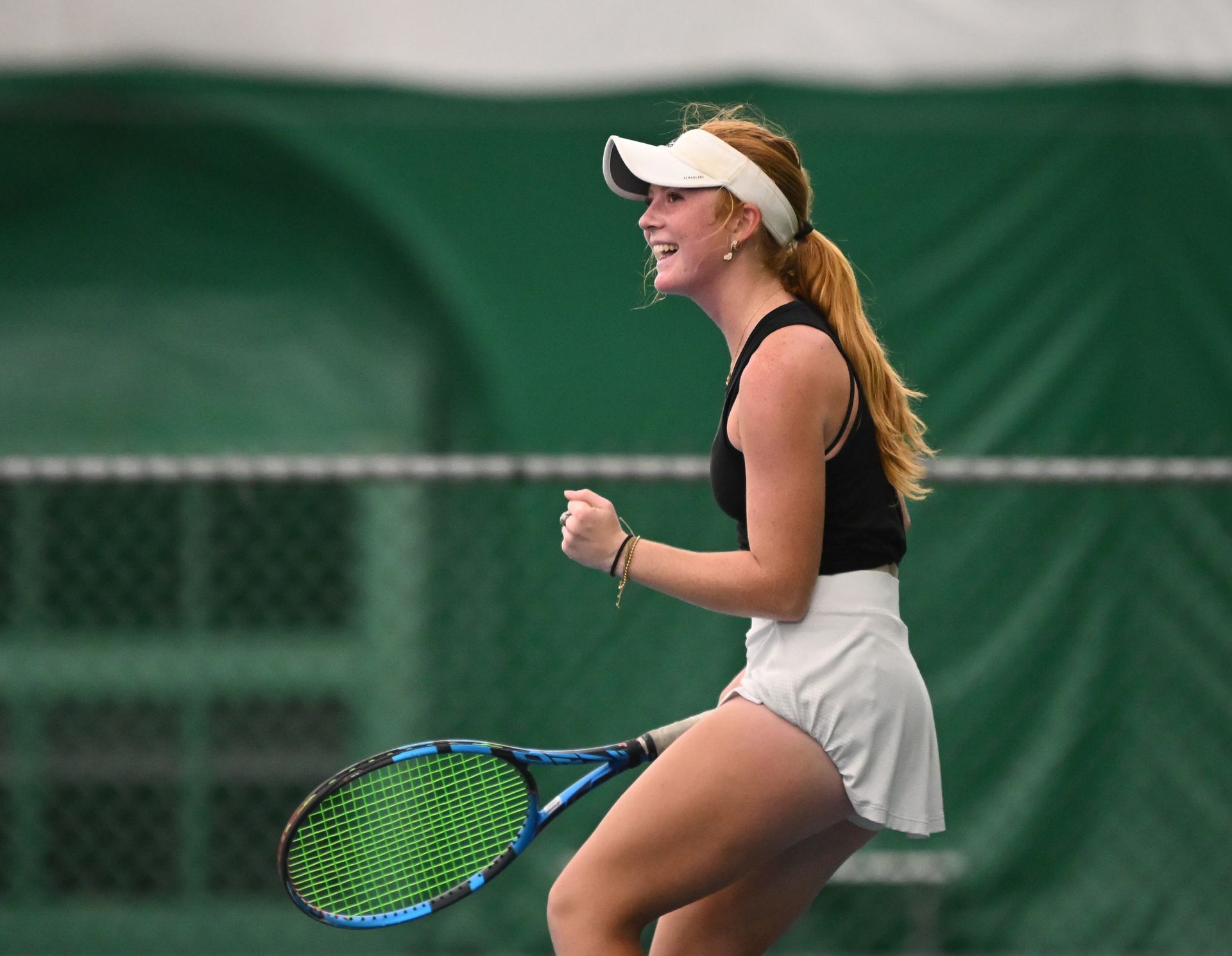 Tennis player in a black tank top and white skirt celebrates on court, holding a racket, with a joyful expression.