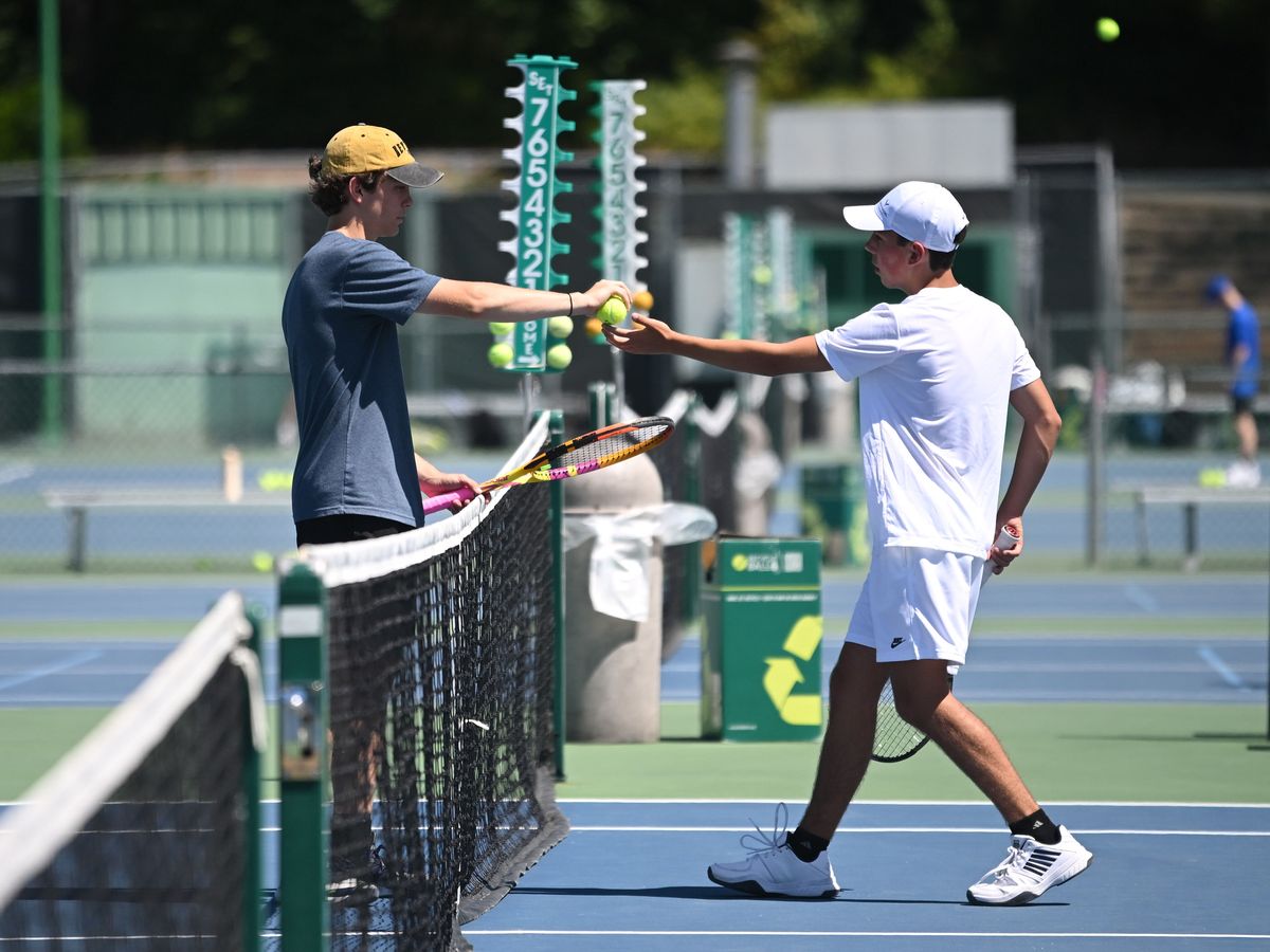 Two tennis players exchange tennis balls over the net on an outdoor court, with one in motion and scoreboard visible in the background.
