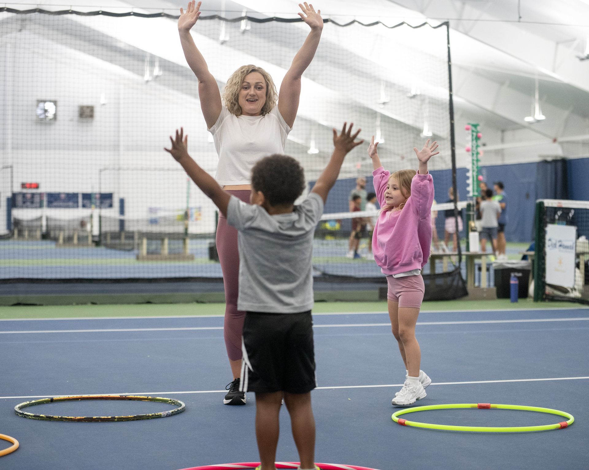 Instructor and two children raise their arms during a playful exercise session on an indoor tennis court with colorful hoops.