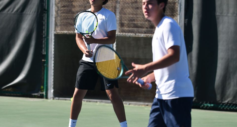 Two high school age kids in white shirts play doubles tennis on an outdoor court, focusing on the ball.