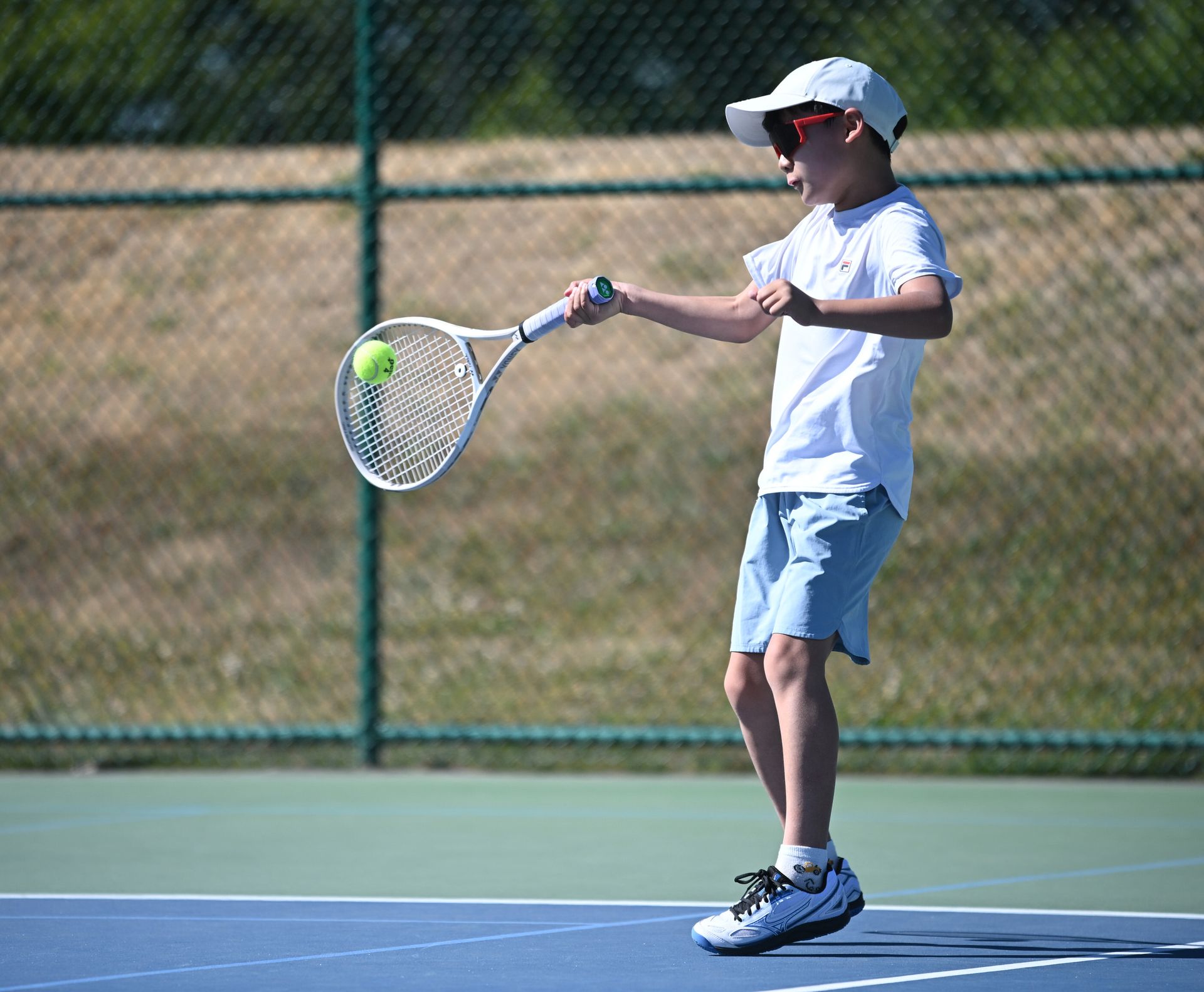 A child in a white cap, sunglasses, and sportswear plays tennis, hitting a ball mid-air on an outdoor court.