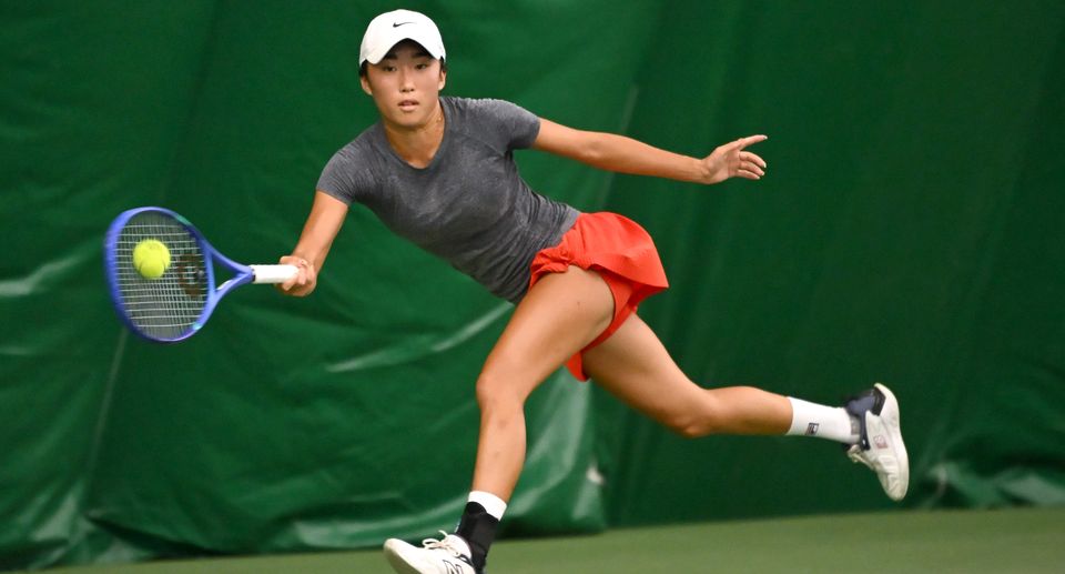 Tennis player in motion, wearing a gray shirt and red skirt, skillfully returns a shot with a forehand swing on an indoor court.