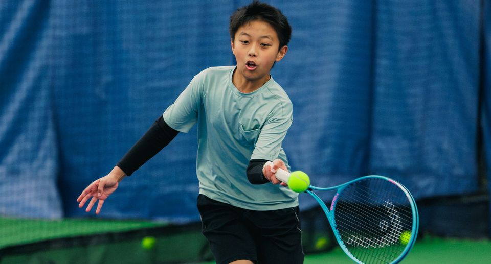 A young player in a light blue shirt and black shorts hits a forehand on an indoor tennis court, with a blue backdrop behind.