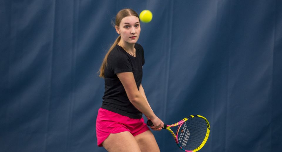 A young tennis player in pink shorts prepares to hit a ball with her racket on an indoor court.