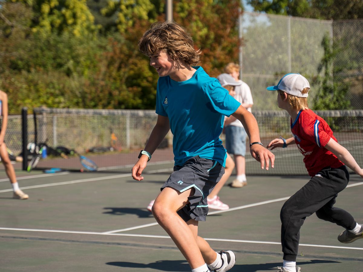 Kids playing sports on an outdoor court; a boy in a blue shirt and another in red run energetically. Sunny day with trees in the background.