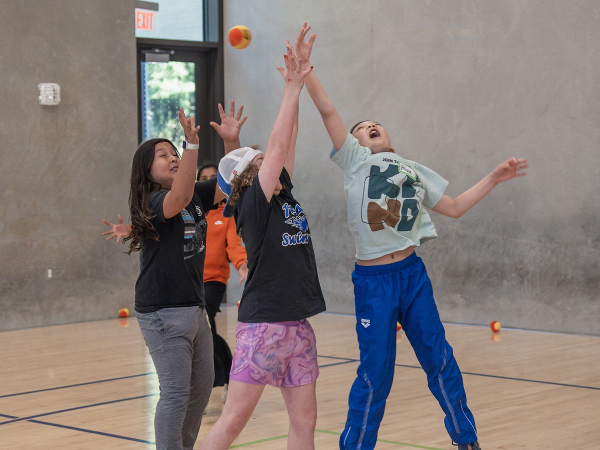Children in a gym jump enthusiastically, reaching for an orange ball in mid-air, showcasing energetic play and teamwork.