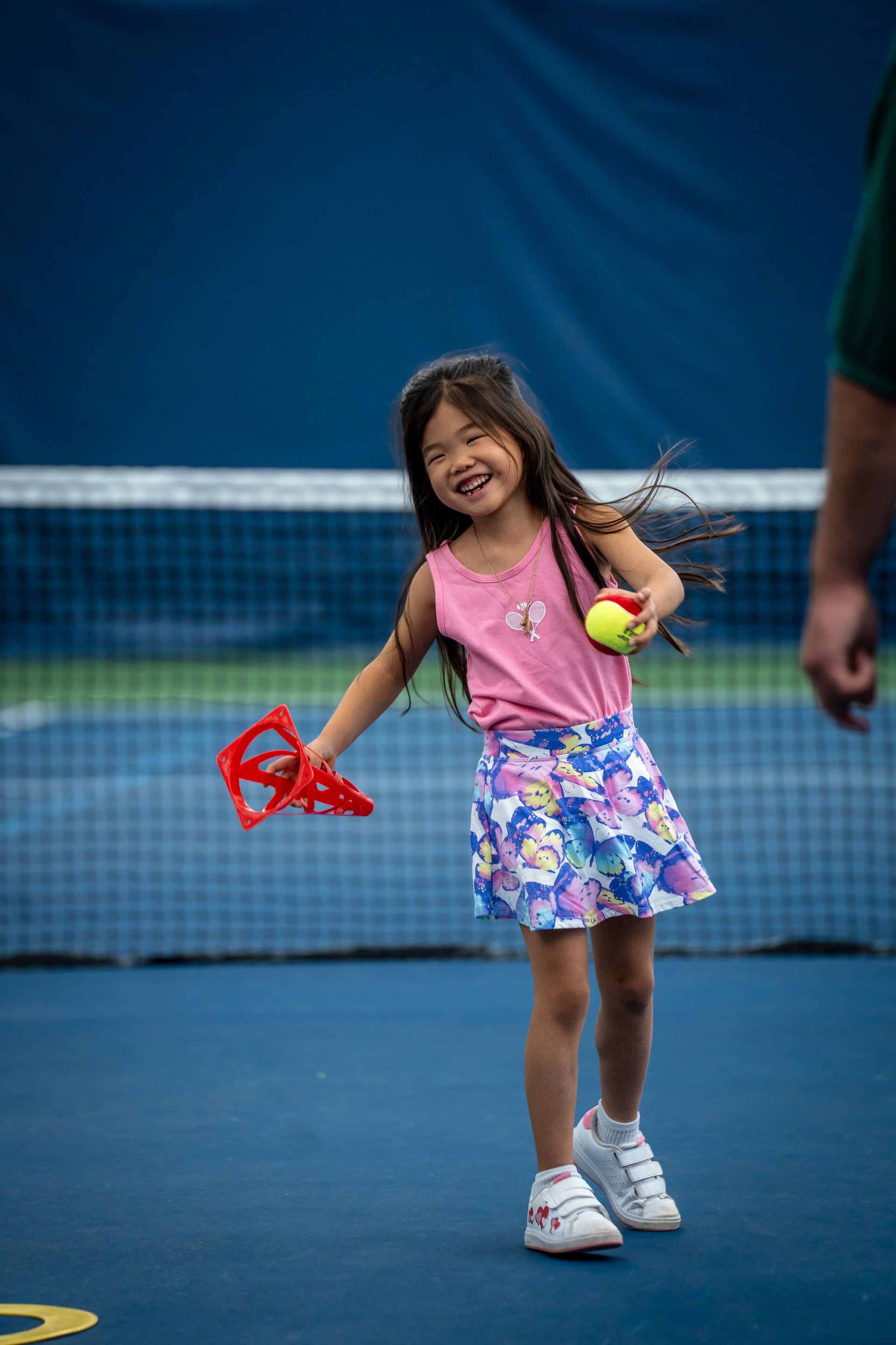 A smiling girl on a tennis court, holding a ball and a red cone, wearing a pink top and a colorful skirt.
