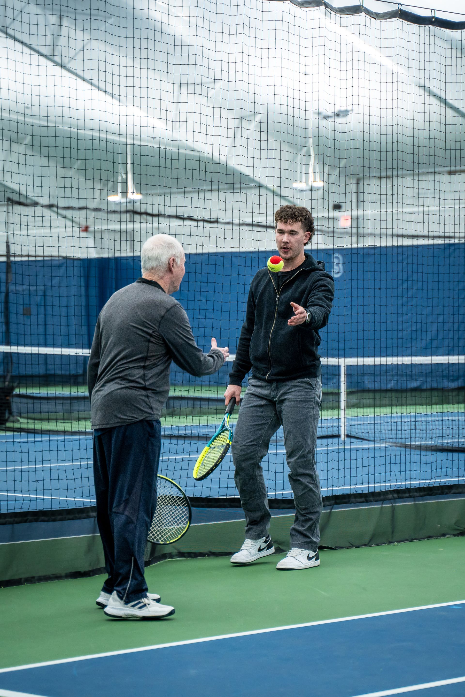 Two men on an indoor tennis court, one tossing a tennis ball to the other, holding a racket, with a net in the background.