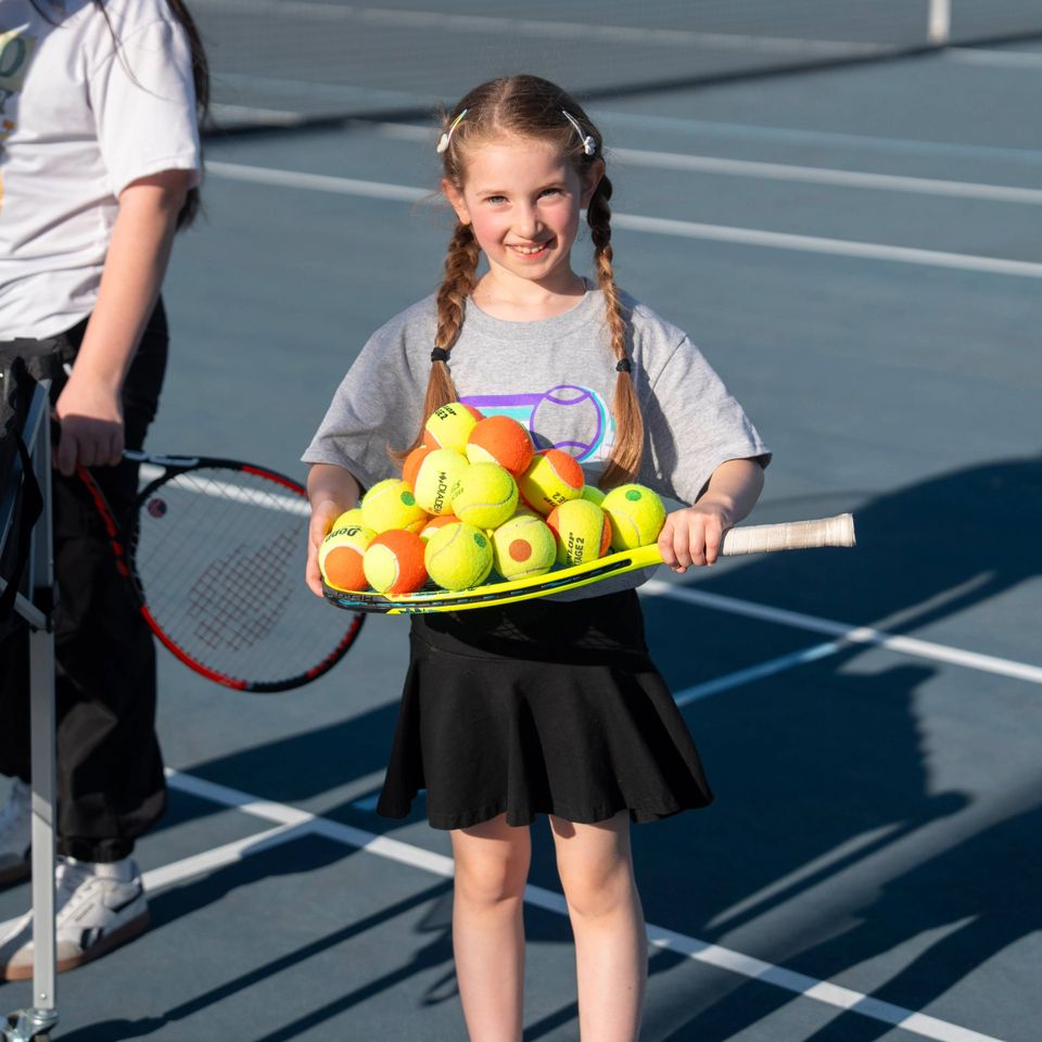 Young girl with braids holds a tennis racket filled with tennis balls on a court. Another person stands nearby with a racket.