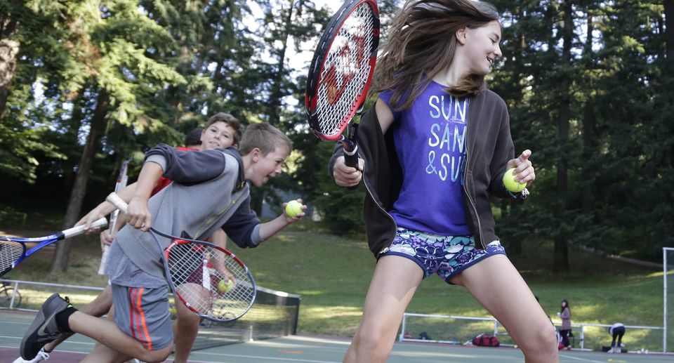 Kids playing enthusiastically with tennis rackets and balls on an outdoor court surrounded by trees.
