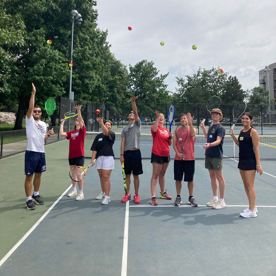 Group of people on a tennis court tossing balls in the air, holding rackets, with green trees and a cloudy sky in the background.