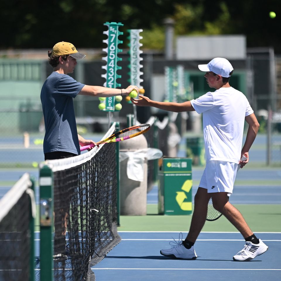 Two tennis players exchange a ball over the net on an outdoor court, with scoreboards visible in the background.