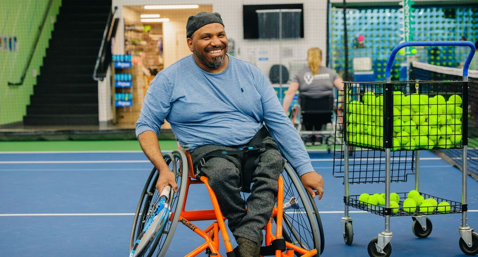 A smiling man in a wheelchair on an indoor tennis court, with a basket of tennis balls nearby, wearing a blue shirt and black cap.