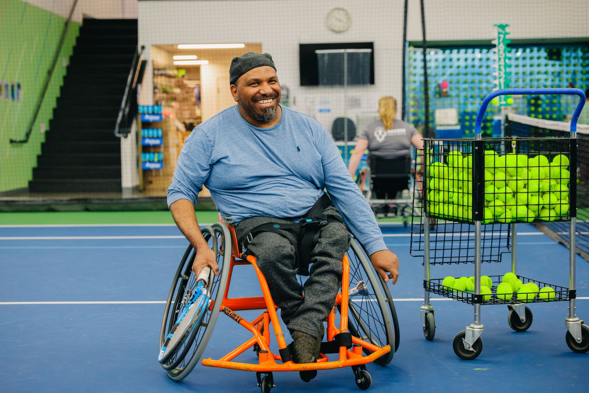 A person in a wheelchair smiles on a tennis court, with a cart of tennis balls nearby.