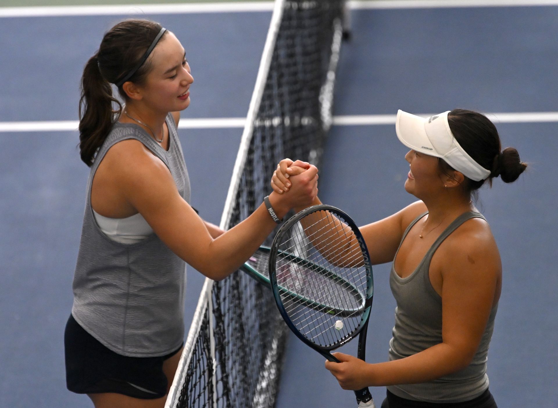 Two tennis players shake hands at the net on an indoor court, both smiling and holding their rackets.