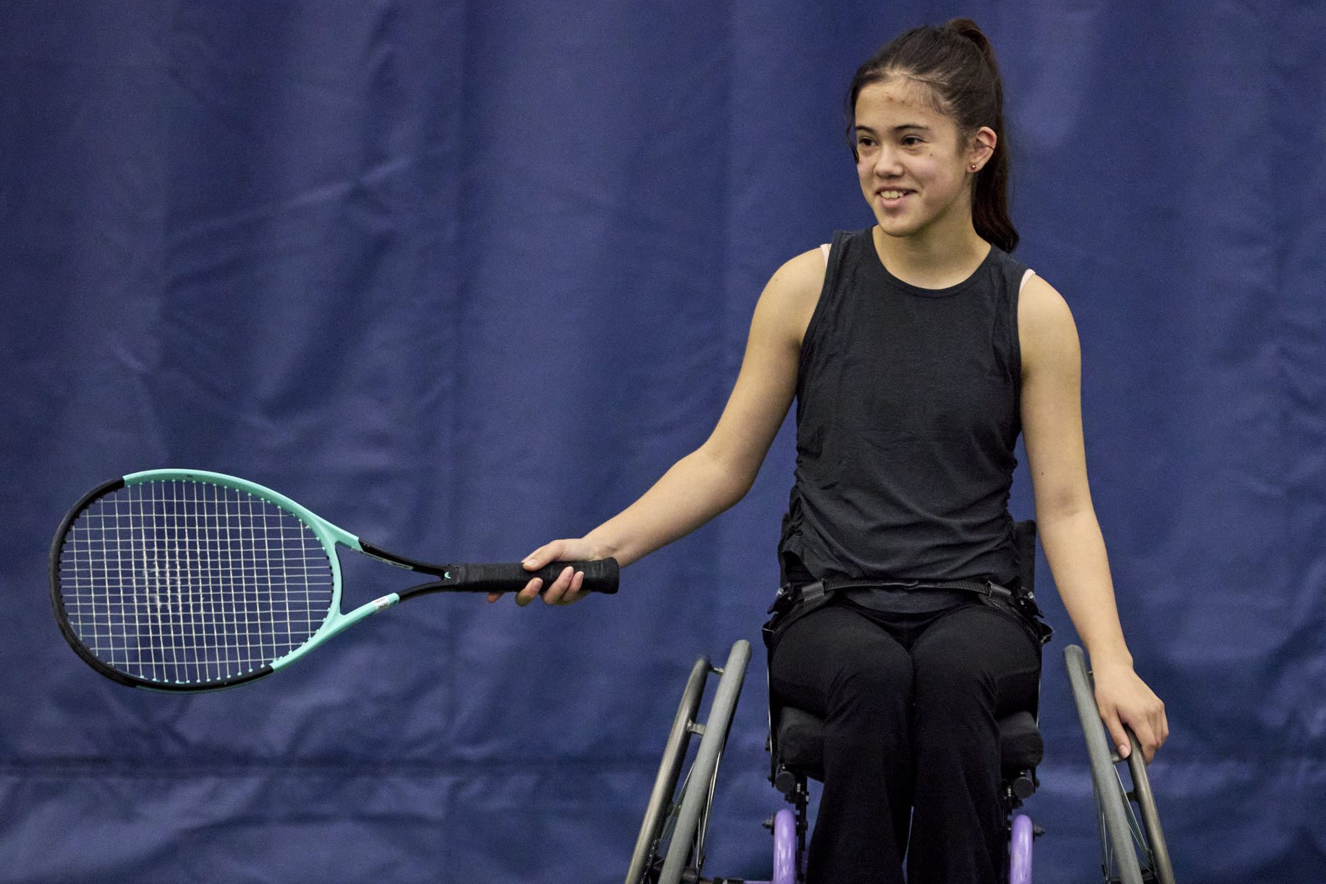 A girl in a wheelchair holds a tennis racket, smiling, with a dark blue curtain background.