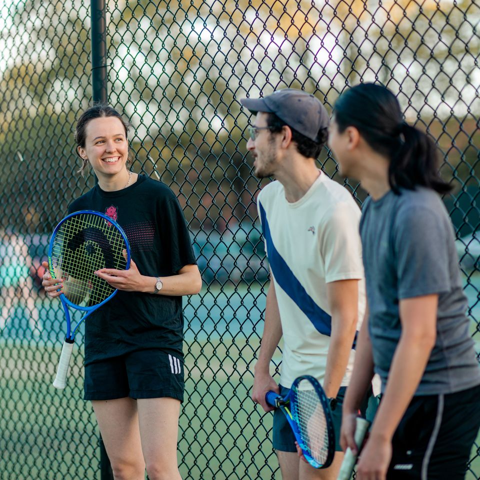 People with tennis rackets chat and smile near a chain-link fence at an outdoor court.