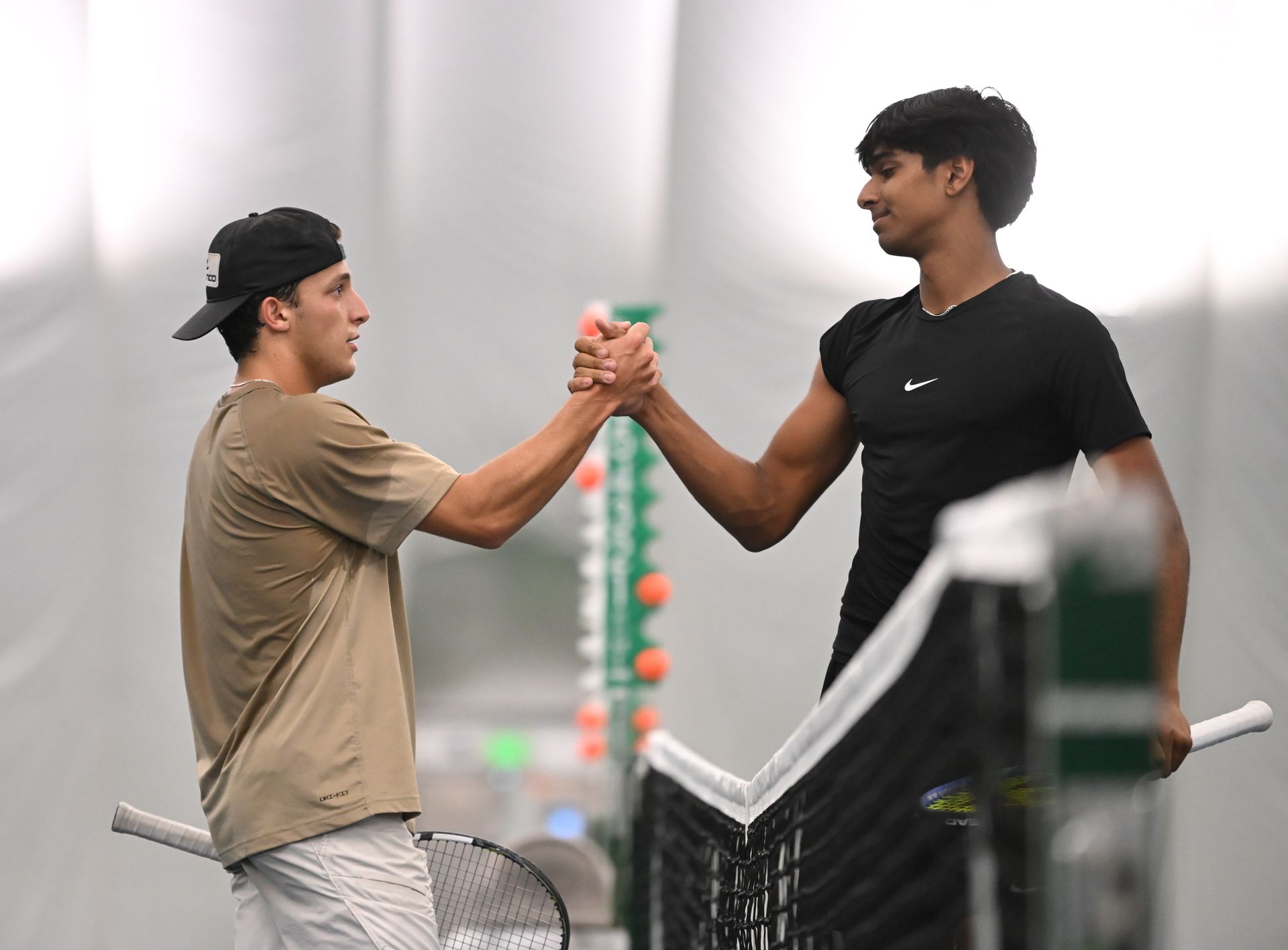Two tennis players exchange a handshake over the net on an indoor court, holding their rackets in their other hands.