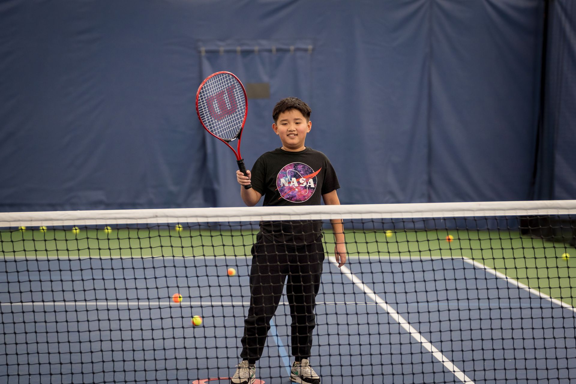 A Love Serving Autism player stands on a tennis court, holding a racket and wearing a NASA shirt, with scattered tennis balls around them.