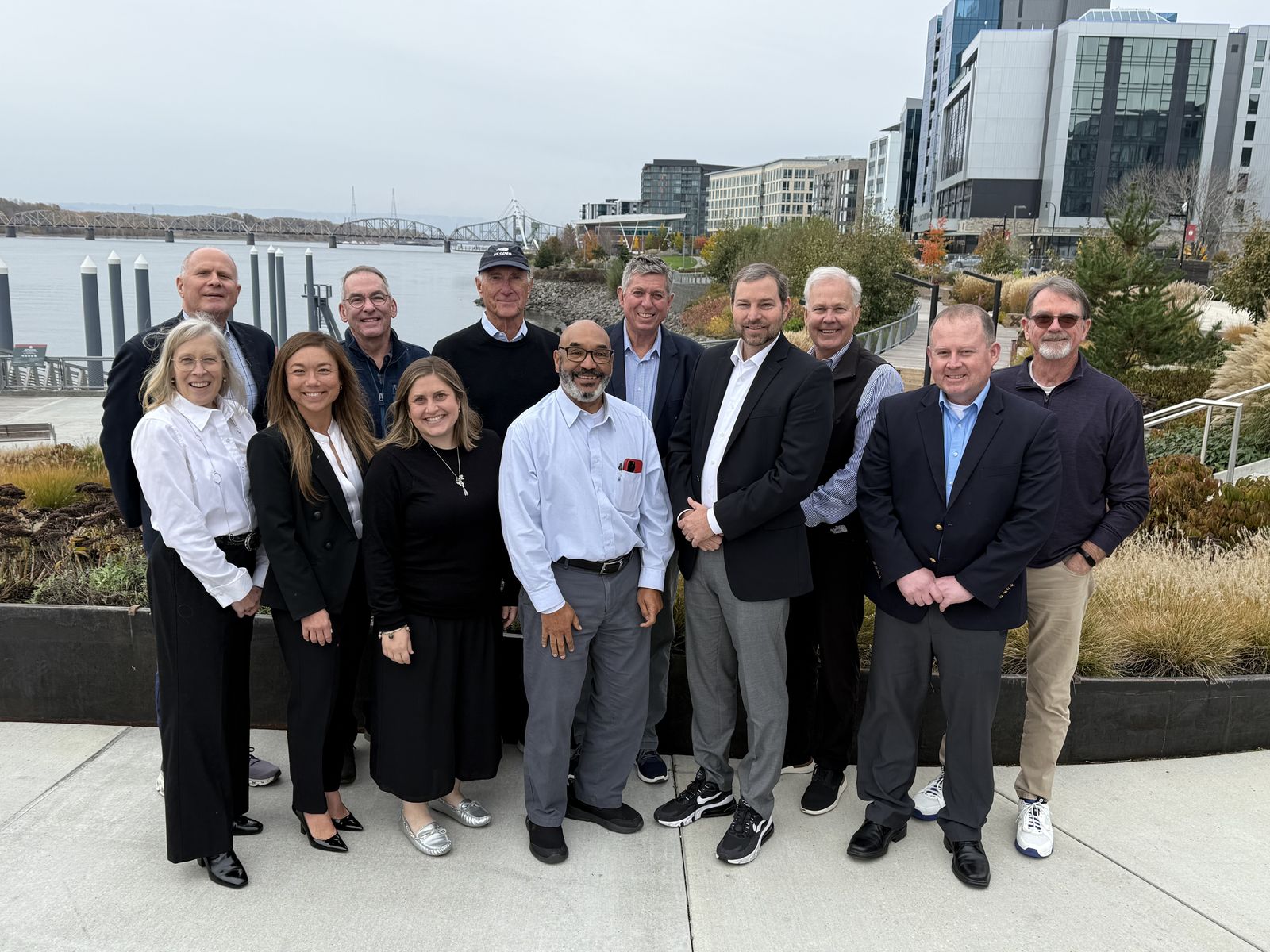 Group of 13 people smiling, standing outdoors near a waterfront with modern buildings in the background on a cloudy day.