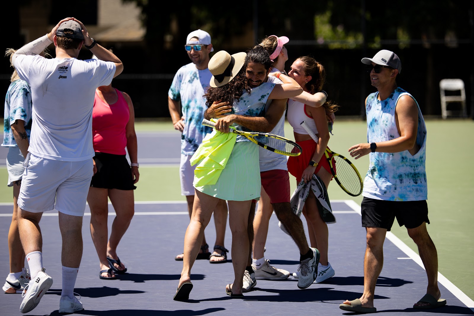 A group of people on a tennis court celebrate joyfully, hugging and smiling, with one holding a racquet. Bright sunny day and casual attire.