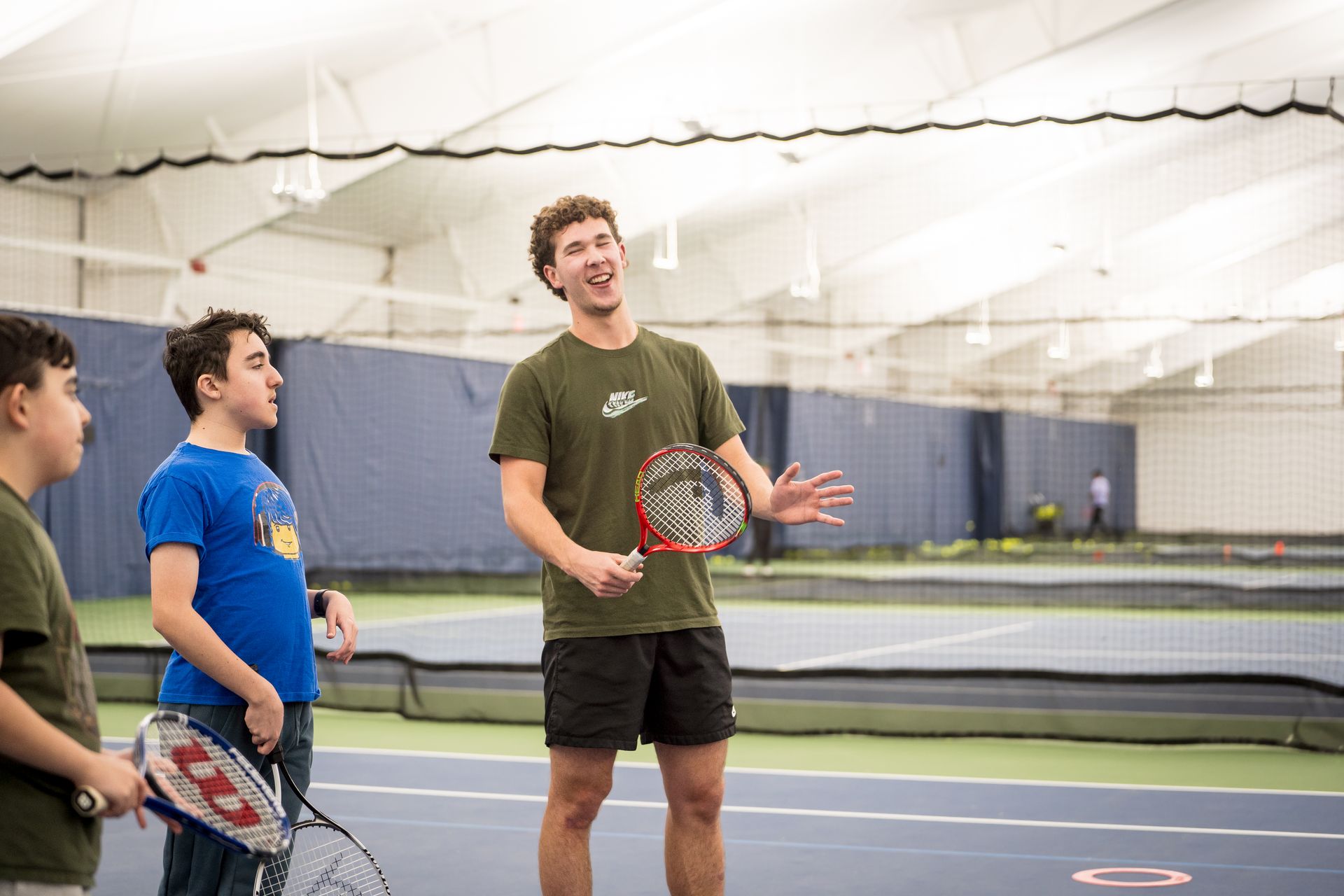 A coach energetically instructs a group of young players on an indoor tennis court, holding a tennis racket.