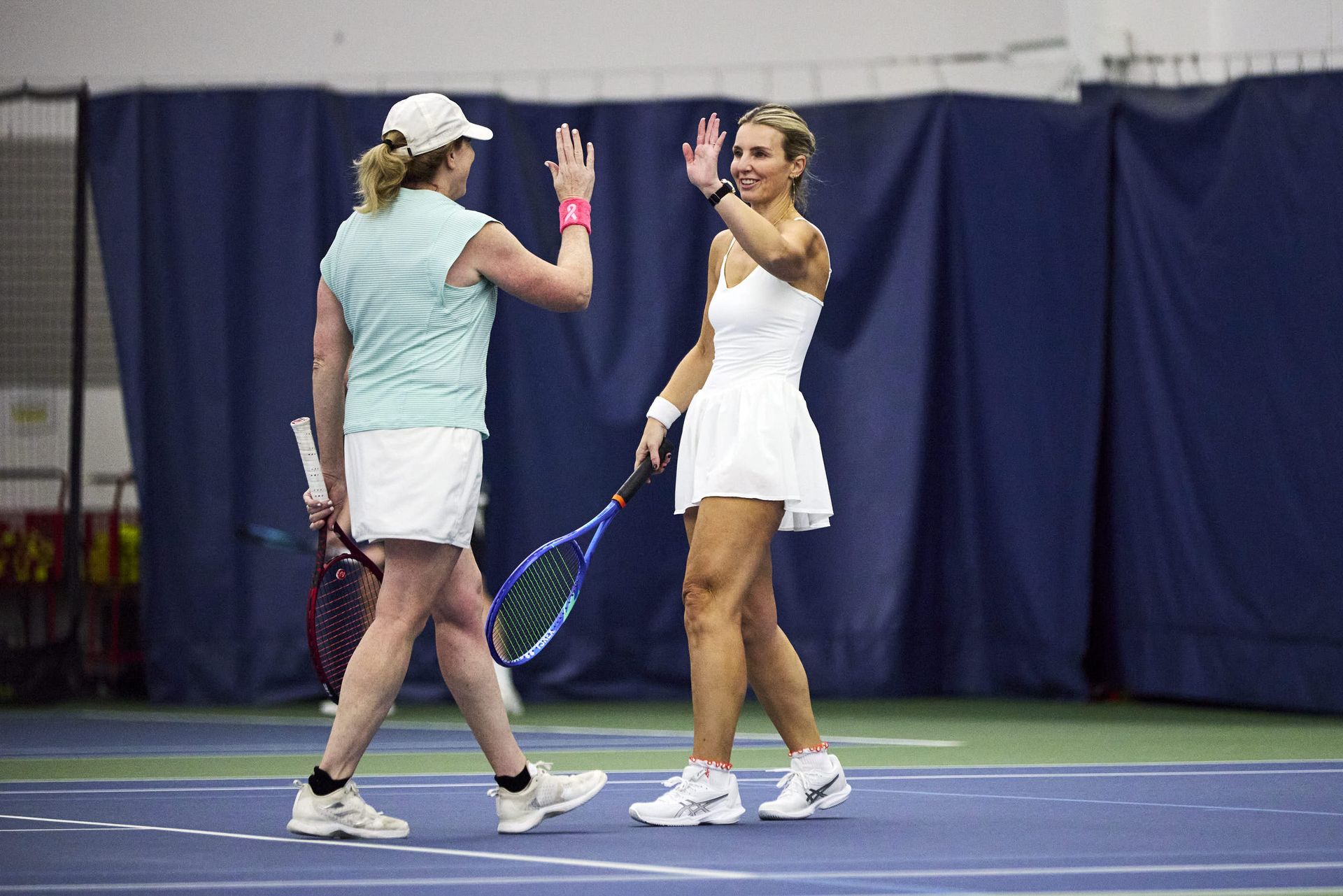 Two female tennis players high-five on an indoor court, celebrating a point. Both hold rackets; one wears a white dress, the other a blue tank top.