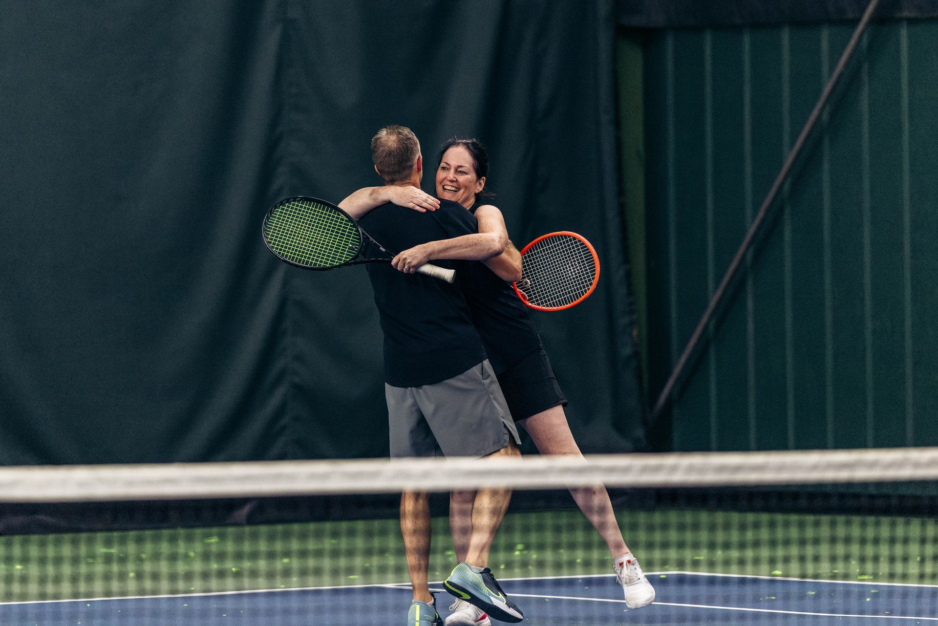 Two people hugging joyfully on an indoor tennis court, both holding rackets, with a net visible in the foreground.