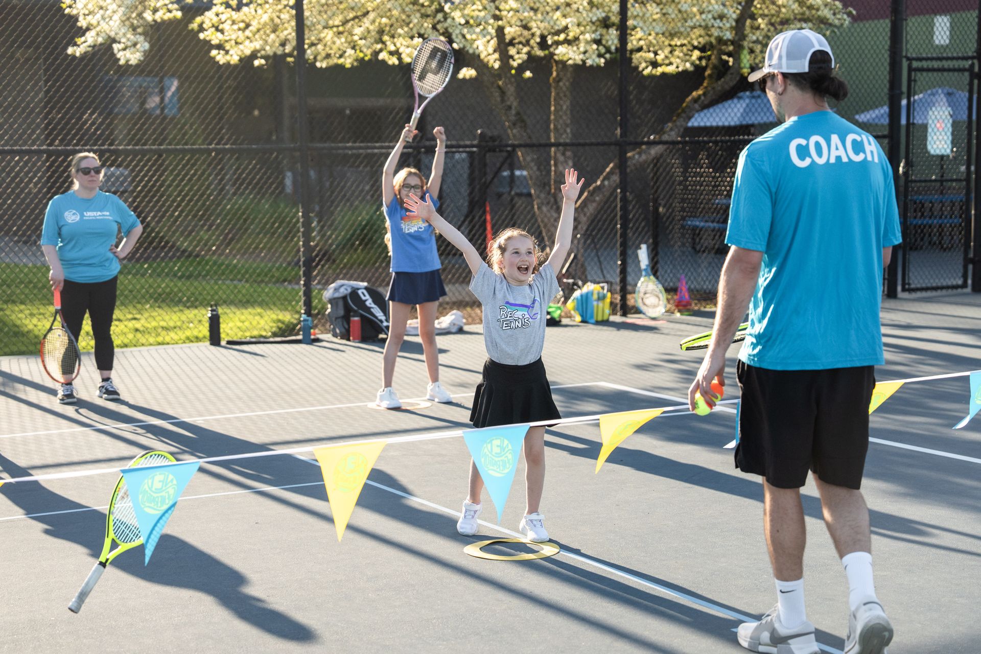 Tennis coach instructing children on a sunny court with colorful buntings and rackets, while a young girl raises her arms excitedly.