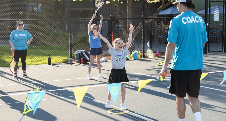 Tennis coach instructing children on a sunny court with colorful buntings and rackets, while a young girl raises her arms excitedly.