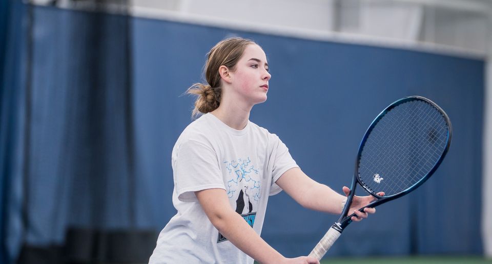 A woman on an indoor tennis court holding a racket, preparing to return a ball.