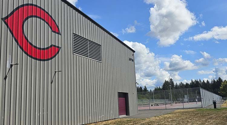 Metal building with a large red "C" logo on the side, near a tennis court under a partly cloudy sky.